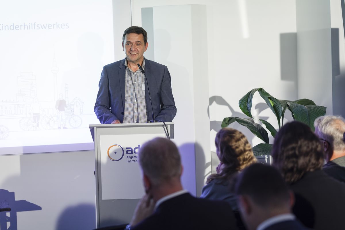 Holger Hofmann Holger Hofmann, Bundesgeschäftsführer des Deutschen Kinderhilfswerkes, am Podium. Im Hintergrund eine Leinwand, auf die Licht projiziert wird.